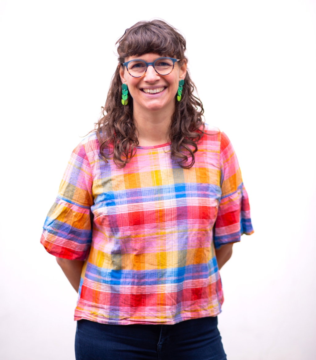 Headshot of Sophie wearing a colourful top, green earrings and glasses, against a white backdrop