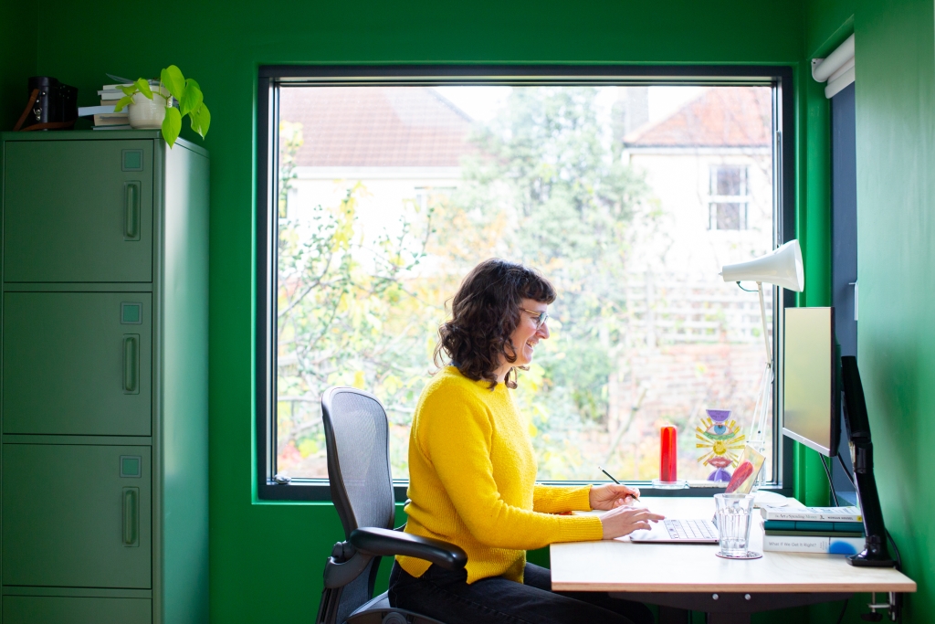 Sophie sitting at a desk wearing a yellow jumper looking at a computer with a large picture window in the background